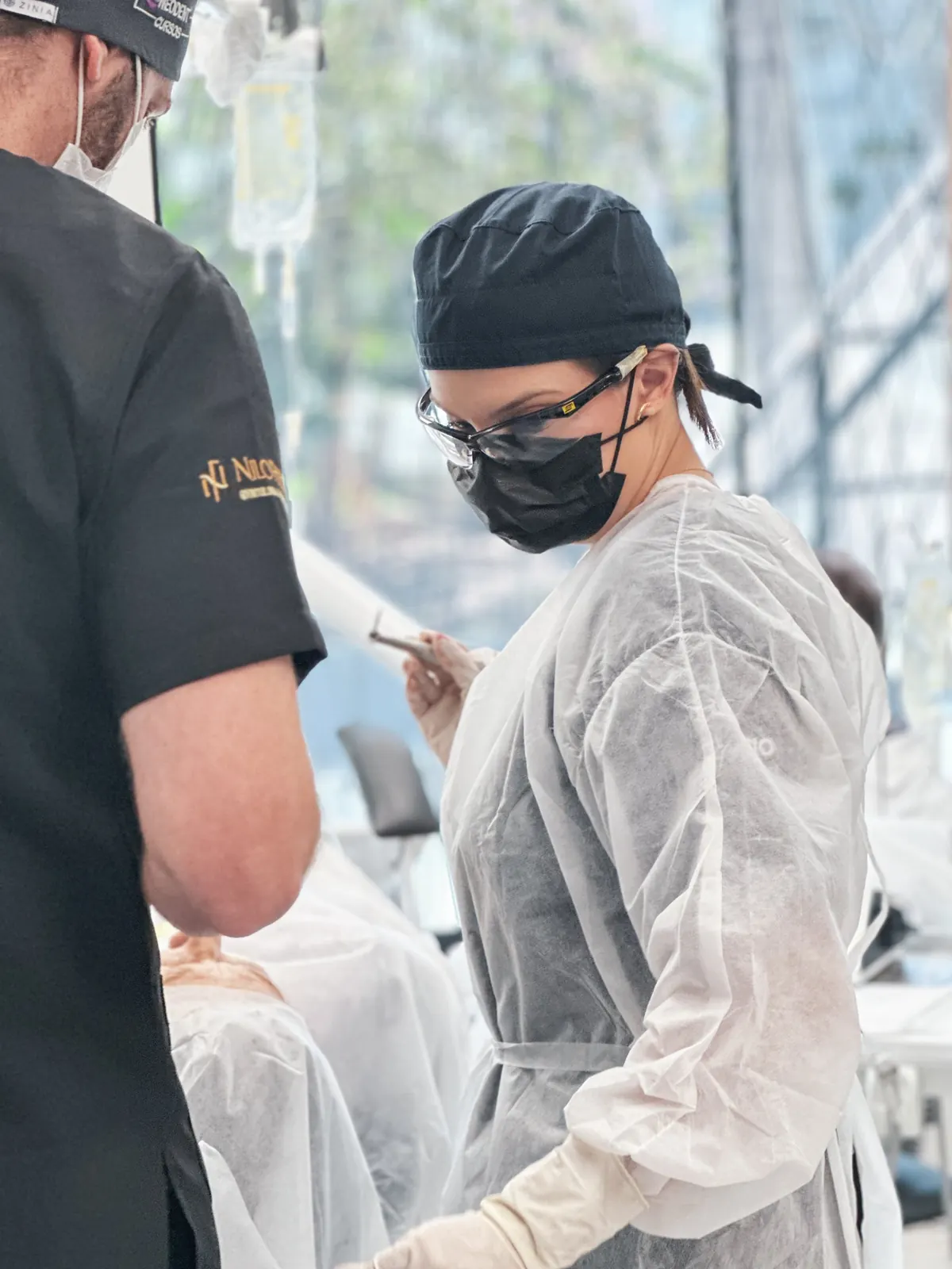 Profissional de avental descartável, touca e óculos de proteção auxiliando um procedimento